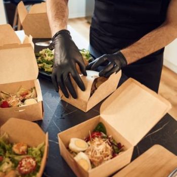 View from the side of meal, vegetables. Man is packing food into the paper eco boxes. Indoors, restaurant.
